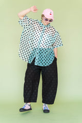 Full-body shot of a model playfully posing in the size-inclusive Nettle Studios Sheer Mint Tulip Top paired with black polka dot trousers. The oversized button-up shirt brings soft structure and playful graphic styling to a warm-weather look.
