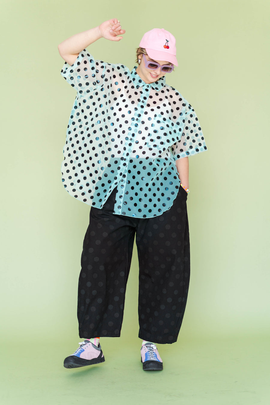 Full-body shot of a model playfully posing in the size-inclusive Nettle Studios Sheer Mint Tulip Top paired with black polka dot trousers. The oversized button-up shirt brings soft structure and playful graphic styling to a warm-weather look.