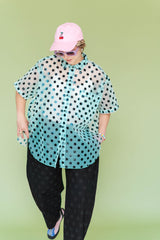 Mid-shot of a model standing in the size-inclusive Nettle Studios Tulip Top, looking down while showcasing the dramatic curved petal hemline for easy pocket access. The sheer green blouse features a black polka dot pattern.