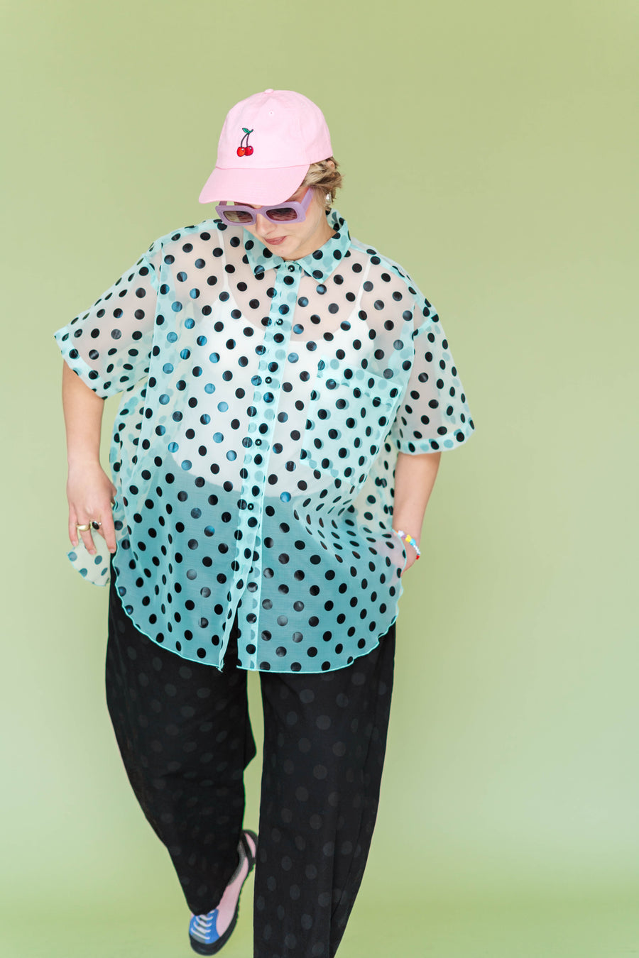 Mid-shot of a model standing in the size-inclusive Nettle Studios Tulip Top, looking down while showcasing the dramatic curved petal hemline for easy pocket access. The sheer green blouse features a black polka dot pattern.