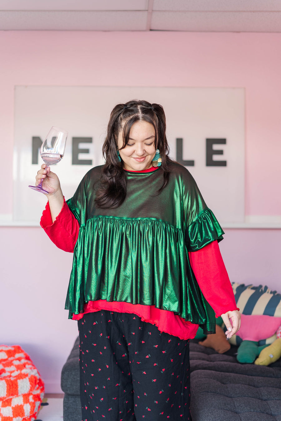 Model smiling while holding a glass, wearing the metallic Green Tinsel Mushroom Top layered over a red long-sleeve top.