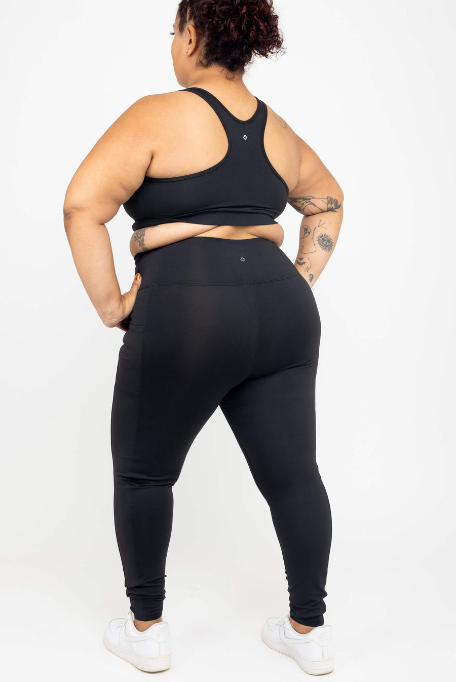 Rear full-body view of a model with arm tattoos wearing black SuperHold compression ankle leggings and a matching racerback sports bra.