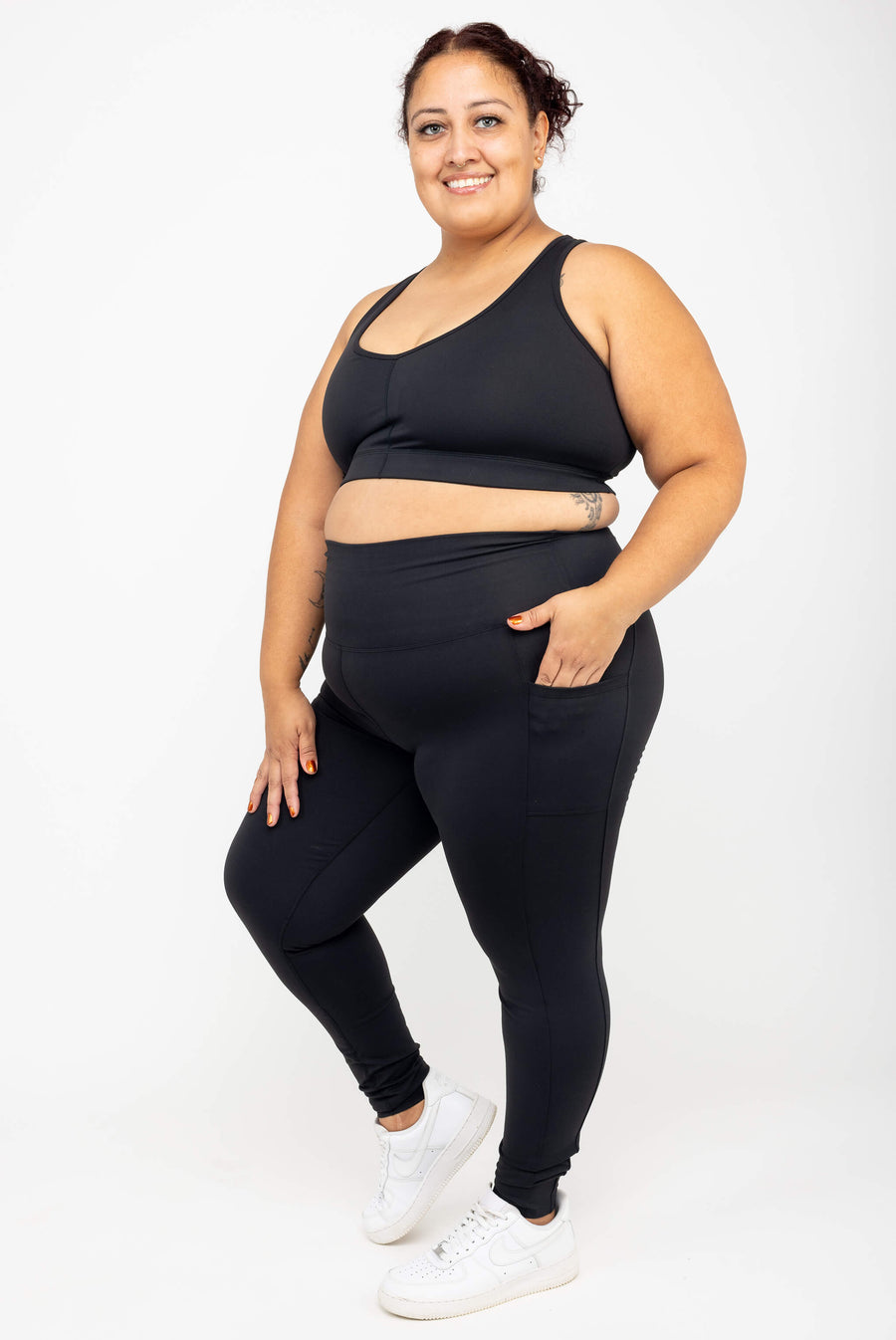 Full-body shot of a brunette model posing with a hand in her pocket wearing black SuperHold ankle leggings and white sneakers.