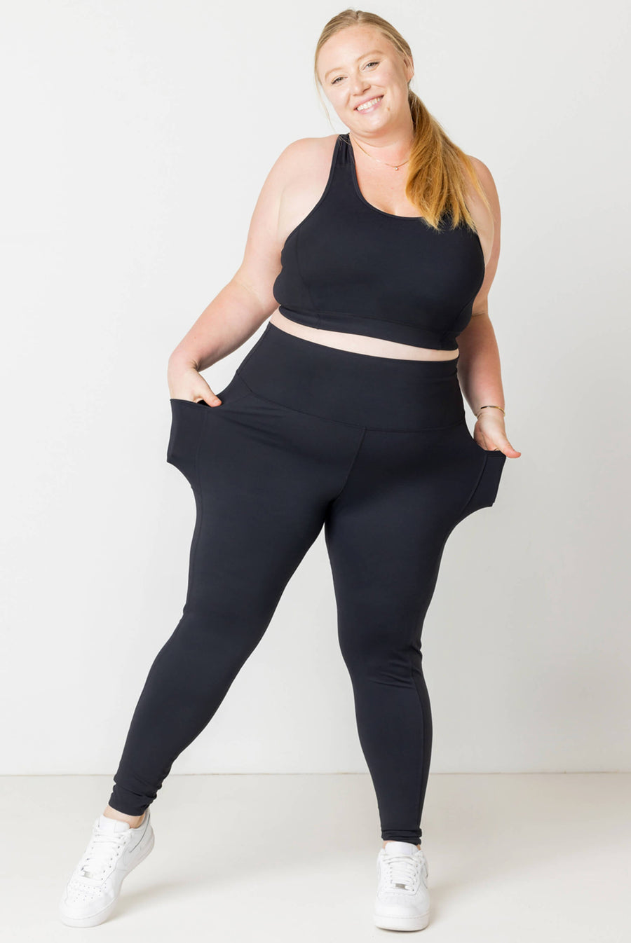 Full-length front view of a smiling blonde model pulling out the side pockets of high-waisted black SuperHold ankle leggings paired with a matching sports bra.