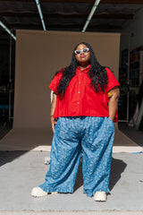 Full-length studio portrait of model Keda wearing the blue floral chambray ruffle pants paired with a bright red short-sleeve button-down shirt and white sunglasses.