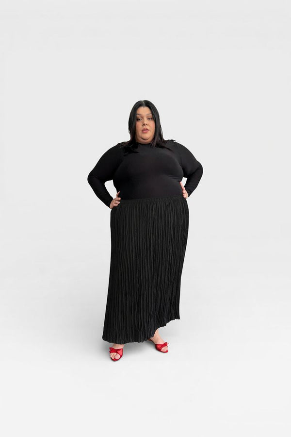 Full-length portrait of a model wearing a size 5 Baacal Rue Pleated Skirt in black paired with a long-sleeve black top and red bow heel sandals.