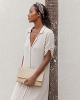 A model leans against a palm tree outdoors, styling a natural straw handwoven envelope clutch with a light cream linen button-up dress.