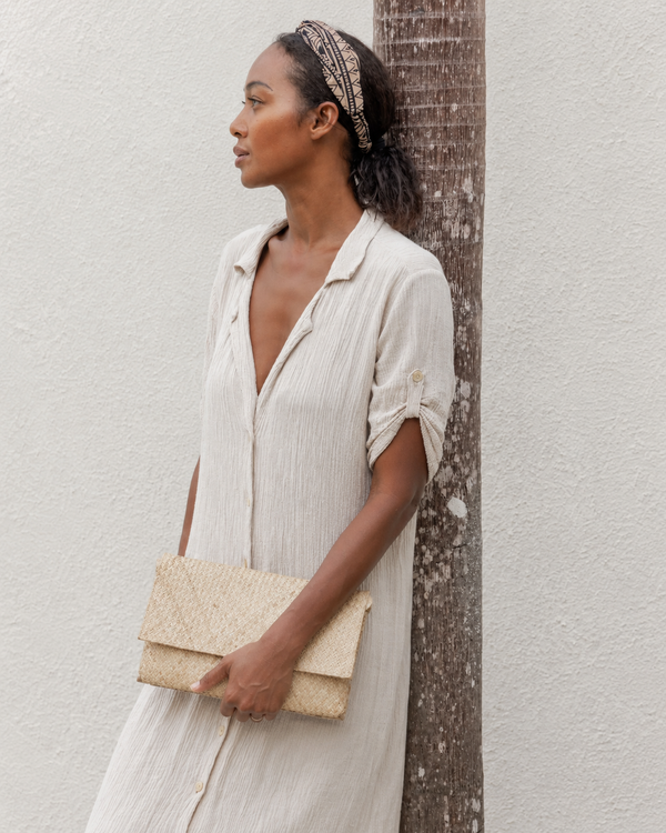 A model leans against a palm tree outdoors, styling a natural straw handwoven envelope clutch with a light cream linen button-up dress.