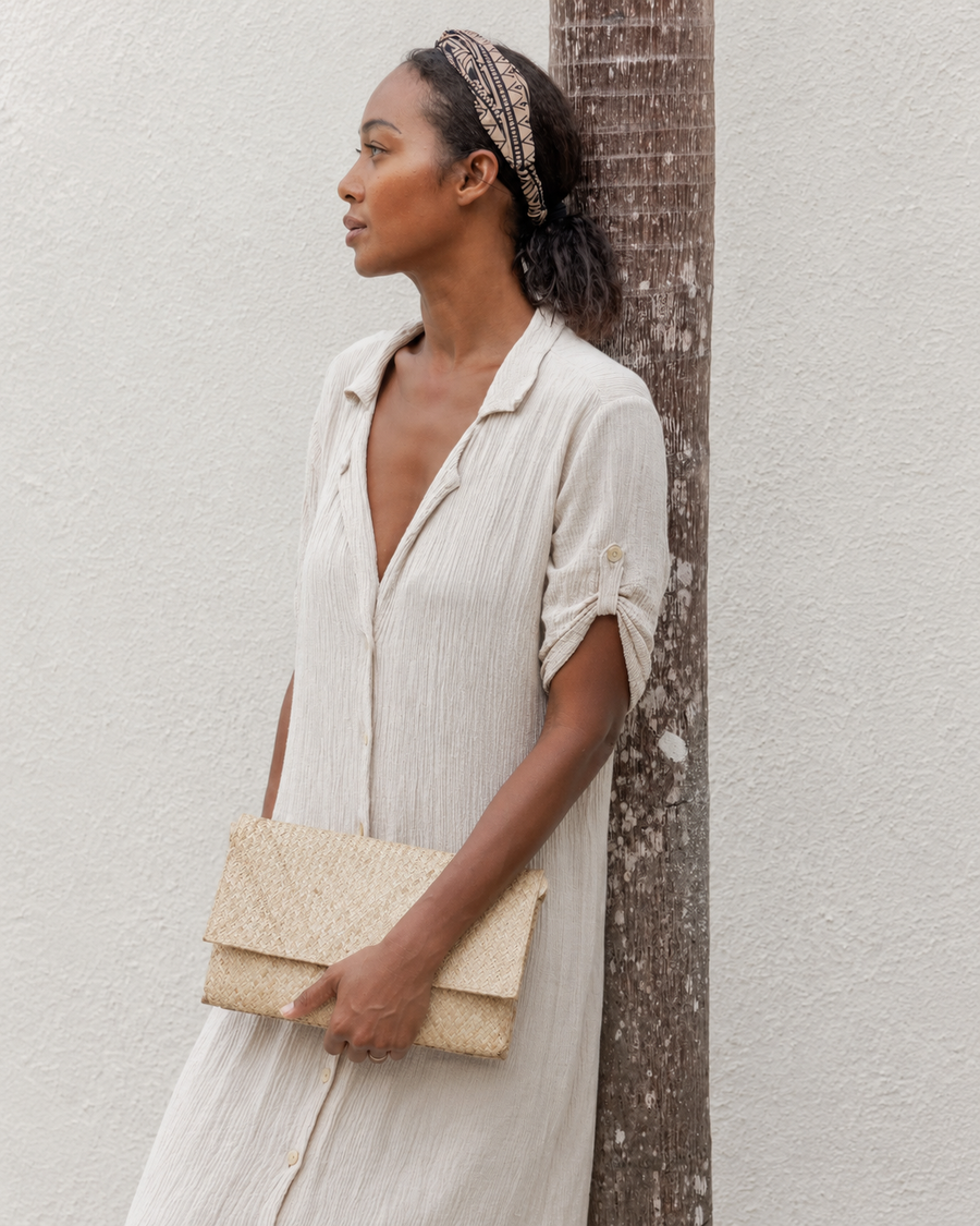 A model leans against a palm tree outdoors, styling a natural straw handwoven envelope clutch with a light cream linen button-up dress.