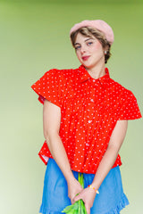 Close-up front view of the size-inclusive Nettle Studios Button Mushroom Top in Cherry Polka Dot. The shot highlights the structured crisp collar, defined chest-seam flare, and short puff sleeves on a model holding flowers against a light green background.