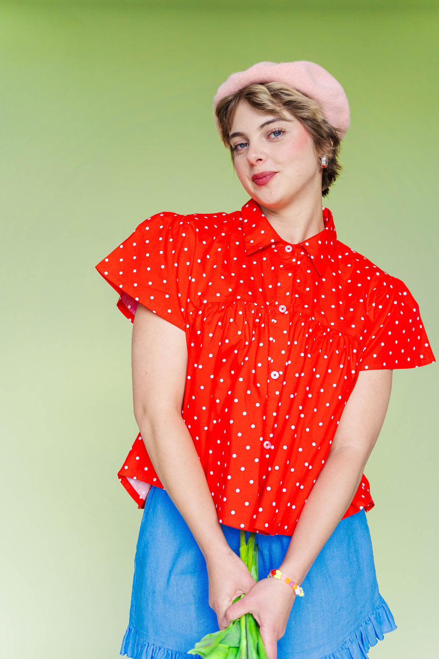 Close-up front view of the size-inclusive Nettle Studios Button Mushroom Top in Cherry Polka Dot. The shot highlights the structured crisp collar, defined chest-seam flare, and short puff sleeves on a model holding flowers against a light green background.