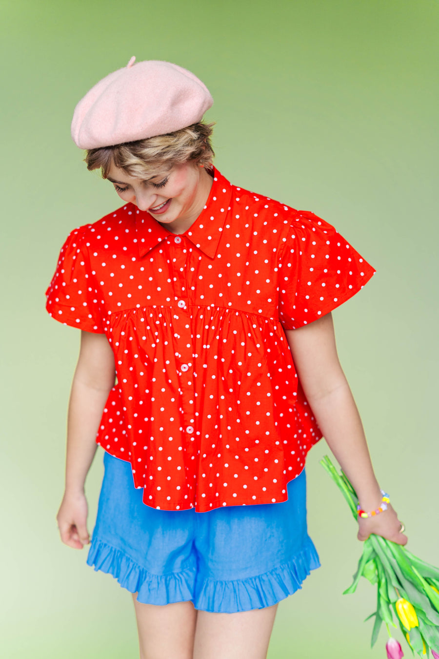 Mid-shot of a model looking down while wearing the Nettle Studios Cherry Polka Dot Button Mushroom Top, showcasing the voluminous, slightly cropped swing shape. Styled with bright blue shorts for a playful, size-inclusive fashion look.