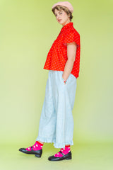 Model wearing Nettle Studios Bleached Floral Chambray Ruffle Pants, a pair of light blue mid-rise wide-leg trousers with an elastic waist and a ruffled hem. Styled in a size-inclusive fashion look against a solid lime green background - Side view