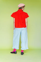 Model wearing Nettle Studios Bleached Floral Chambray Ruffle Pants, a pair of light blue mid-rise wide-leg trousers with an elastic waist and a ruffled hem. Styled in a size-inclusive fashion look against a solid lime green background - Back View. 