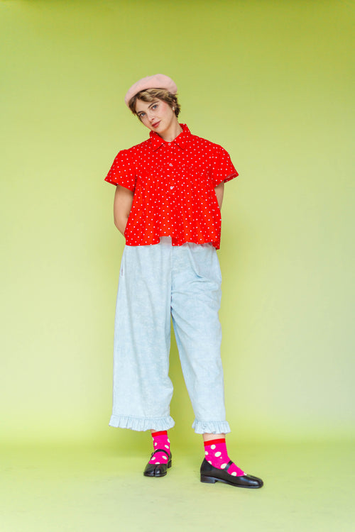 Full-body view of a model in a size-inclusive fit wearing the Nettle Studios Cherry Polka Dot Button Mushroom Top paired with light blue ruffle-hem pants. The red, short-sleeve button-up blouse features white polka dots and a relaxed swing silhouette against a lime green backdrop.