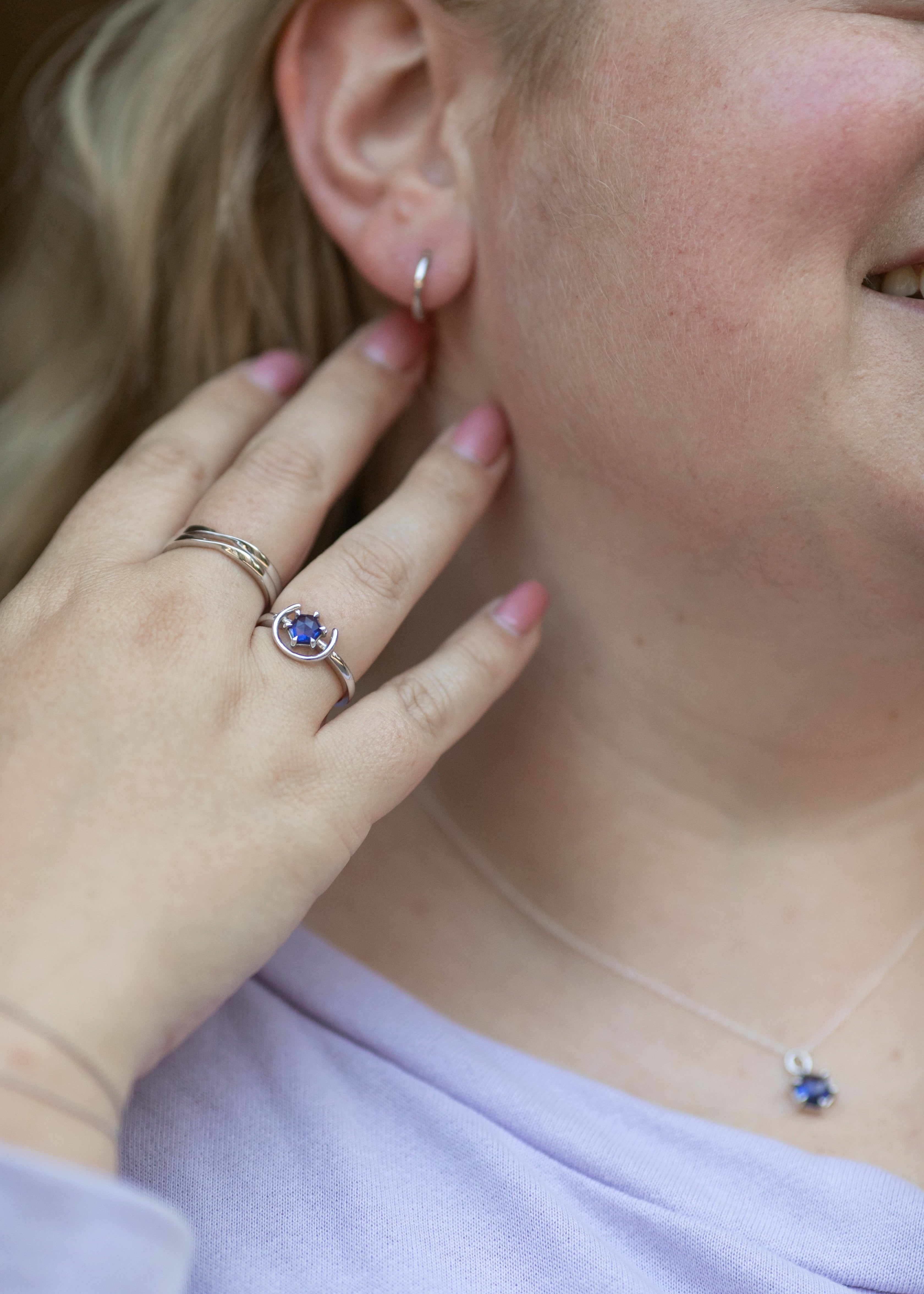 a close up shot of a blonde woman pushing her hair back to show off her simple huggie hoop earring in sterling silver. Her hands feature two twist rings on her middle finger and a gemstone ring in silver and tanzanite on her ring finger. #color_silver