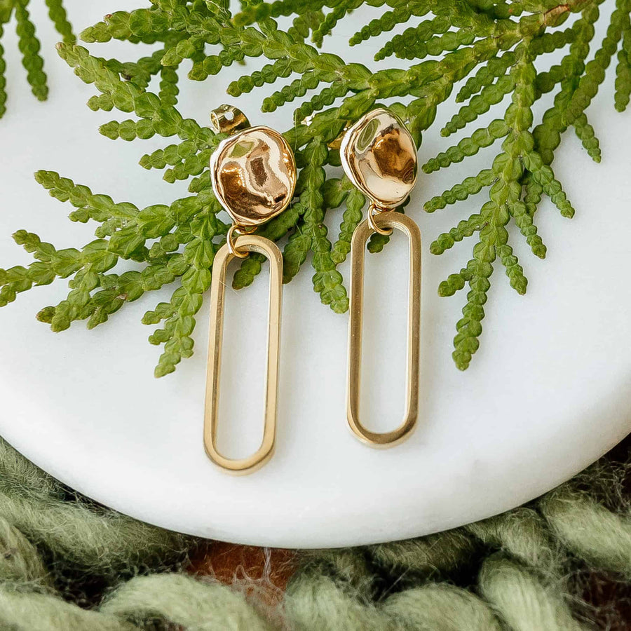Gold earrings on a white surface with green leaves in the background