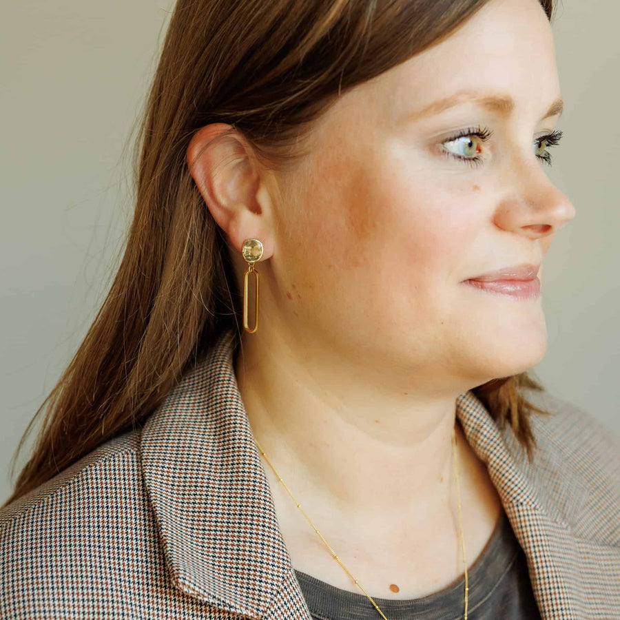 Model wearing the Sela Designs Gold Cleo Earrings, showcasing the 1.5-inch 14k gold-plated oval drop design paired with a plaid blazer.