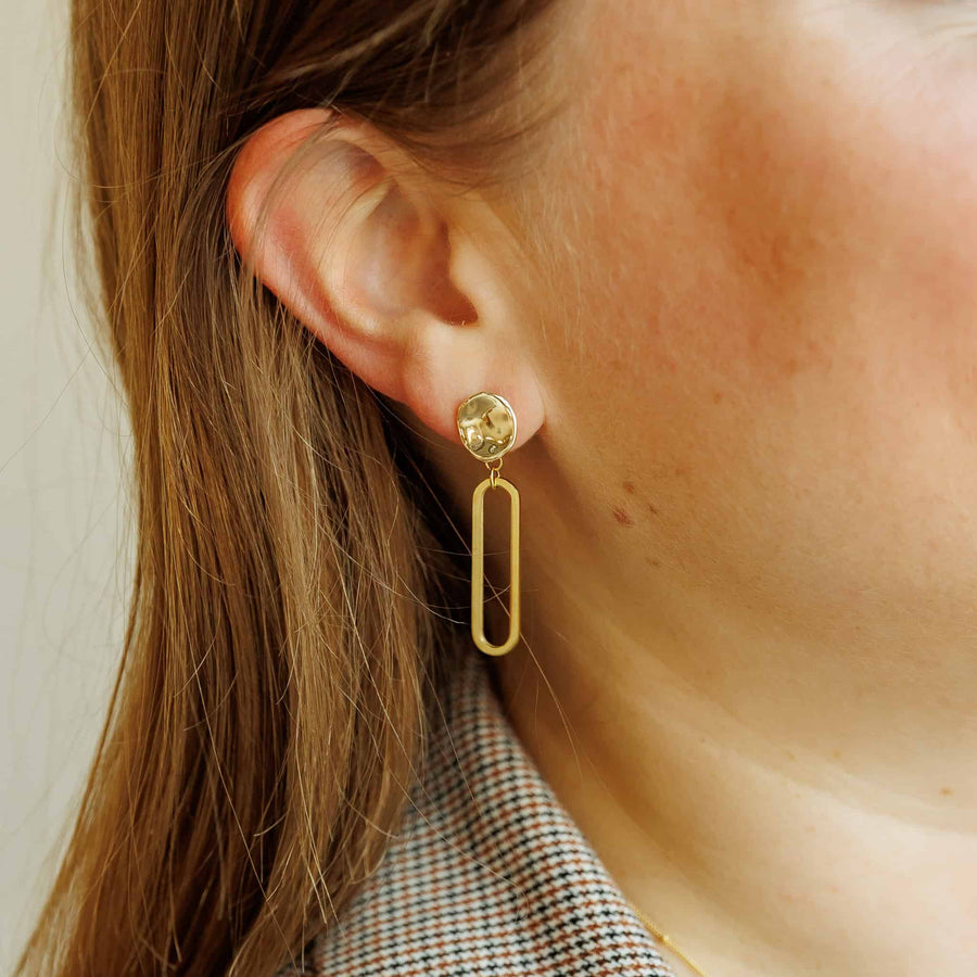 Model wearing the Sela Designs Gold Cleo Earrings, showcasing the 1.5-inch 14k gold-plated oval drop design paired with a plaid blazer.