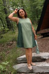 Full-body shot of a tattooed model posing outdoors in the aloe green A-line Beth Dress holding a light blue shoulder bag.