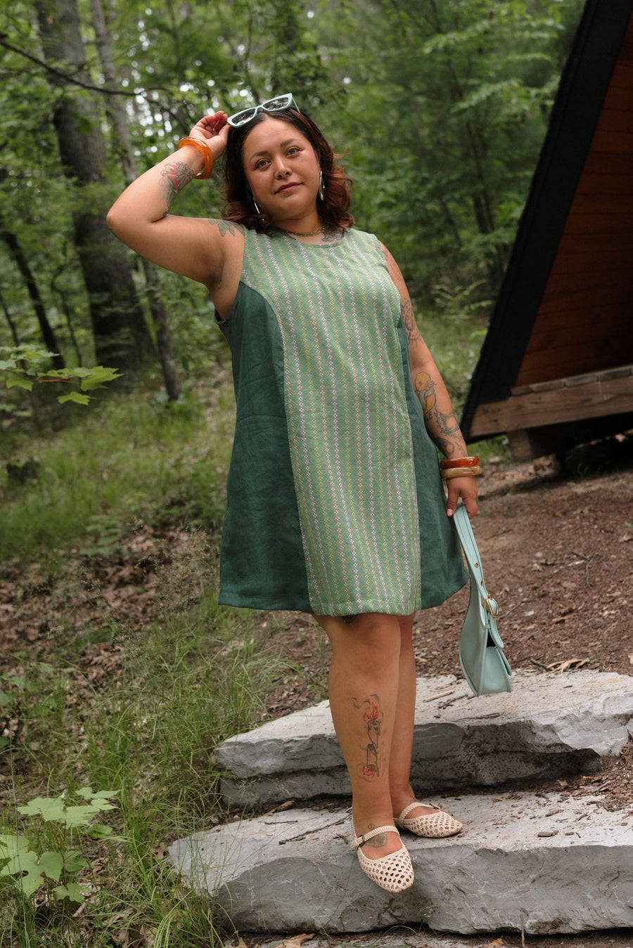 Full-body shot of a tattooed model posing outdoors in the aloe green A-line Beth Dress holding a light blue shoulder bag.