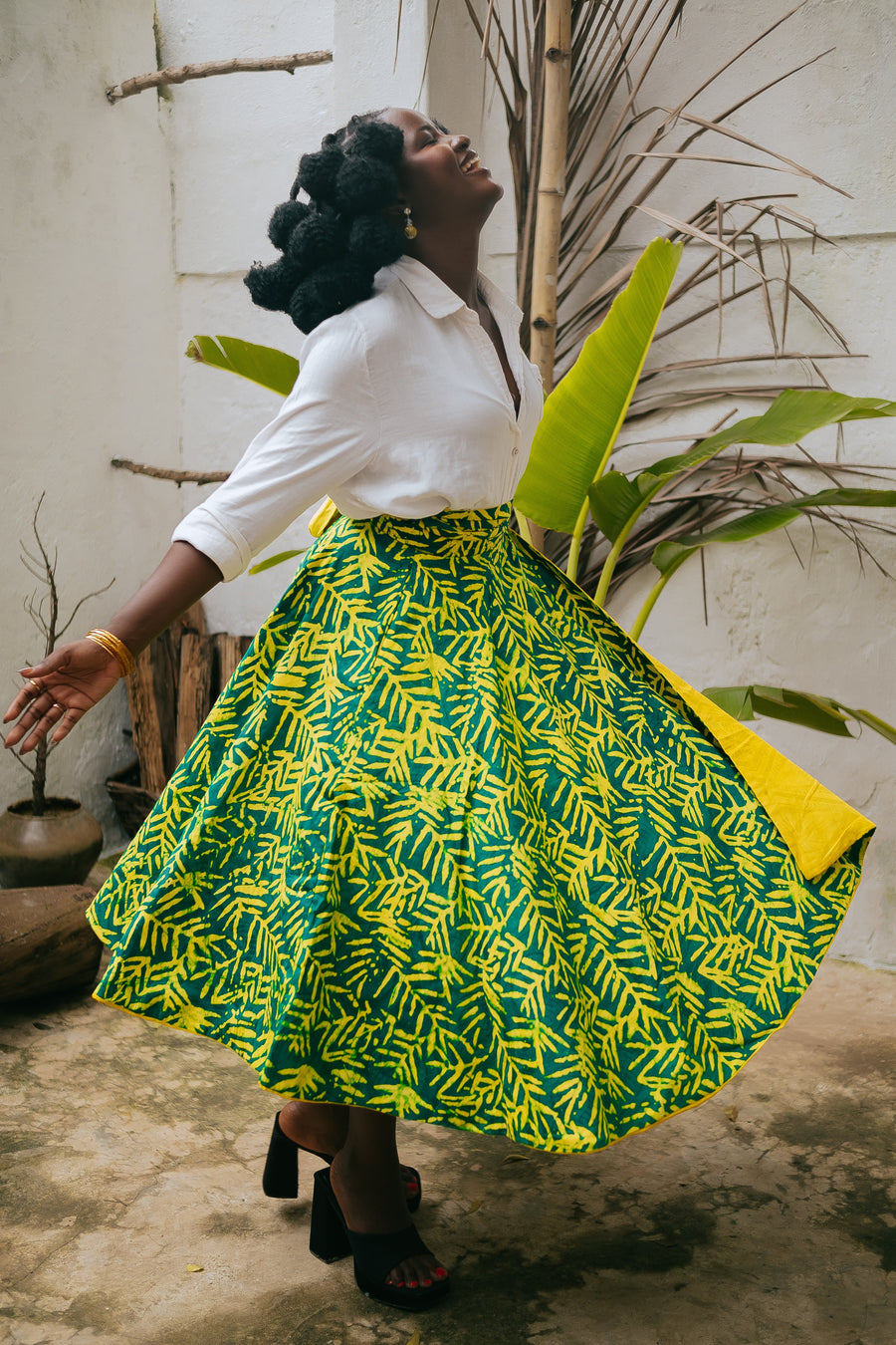 Side profile lifestyle view of a model spinning in the Besida Yellow Palm Reversible Wrap Skirt, capturing its fluid movement and wide flare. The mid-length skirt displays the hand-dyed green and yellow palm-leaf African print with a glimpse of the solid yellow reverse lining. Styled with a white long-sleeve button-down shirt and black block-heel sandals in an outdoor garden setting.