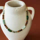 Product display of the Green Phoebe mixed gemstone necklace draped around a textured white ceramic vase.