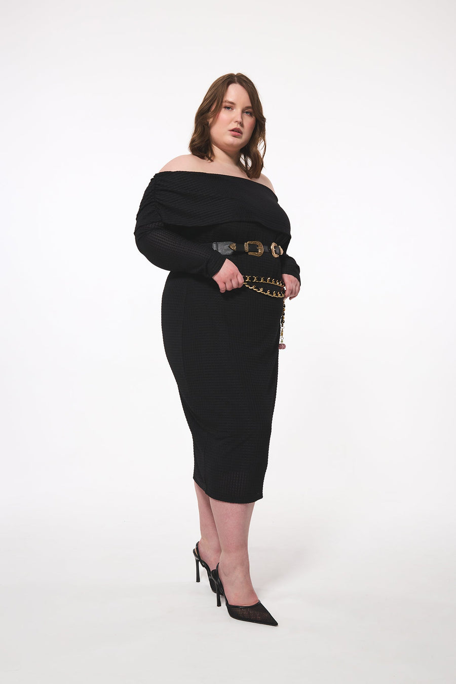 Full-length side profile view of a plus-size model wearing the black geometric knit bodycon midi dress with dual styling belts and black mesh stiletto heels.