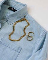 Flat lay product display showing the Parker gold chain necklace arranged on a light blue denim button-down shirt next to gold hoop earrings.