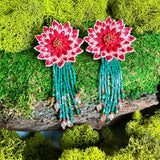 A pair of Coral Red DAHLIA hand-embroidered earrings with green beaded fringe, displayed flat on a textured piece of wood surrounded by bright green natural moss, highlighting the superbloom inspiration behind the design.