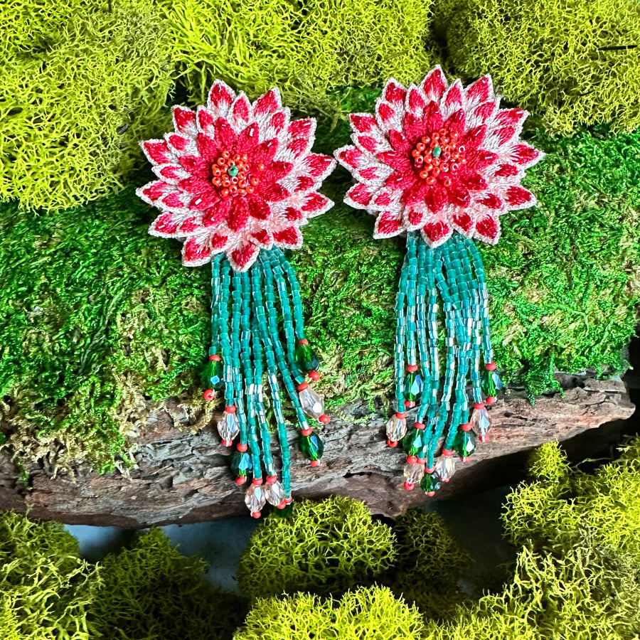 A pair of Coral Red DAHLIA hand-embroidered earrings with green beaded fringe, displayed flat on a textured piece of wood surrounded by bright green natural moss, highlighting the superbloom inspiration behind the design.
