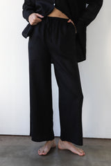 Front view shot of a model wearing the Be Idle wide-leg black linen drawstring pants paired with an open matching black linen shirt.