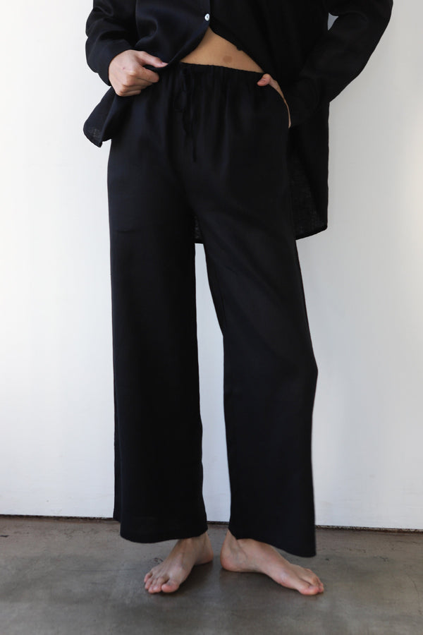 Front view shot of a model wearing the Be Idle wide-leg black linen drawstring pants paired with an open matching black linen shirt.