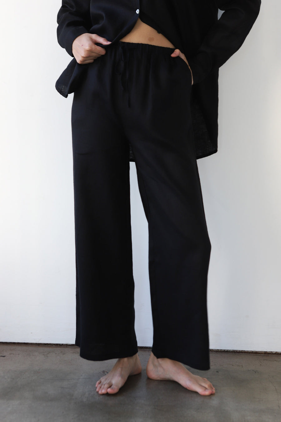 Front view shot of a model wearing the Be Idle wide-leg black linen drawstring pants paired with an open matching black linen shirt.
