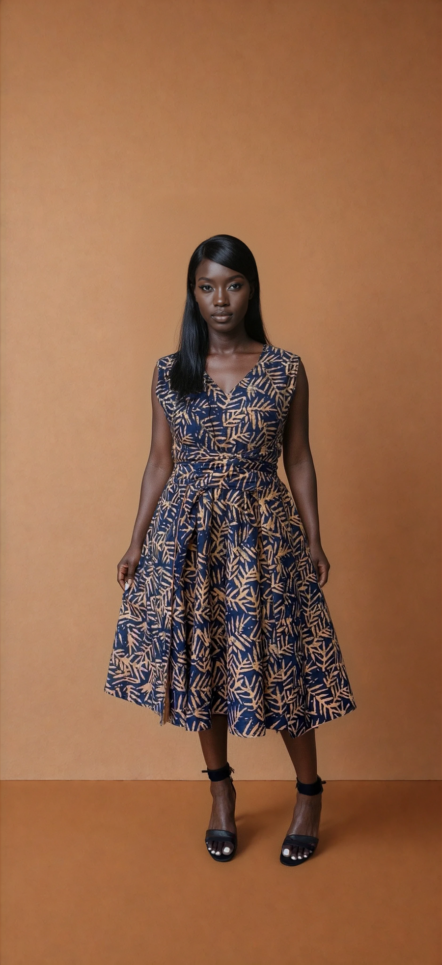 Full-length view of a model wearing the Besida Palm Orange Reversible Dress, showcasing the hand-dyed navy blue and orange-gold palm leaf print side. The sleeveless, V-neck midi dress features a wrapped tie waist and a flowing A-line skirt, paired with black ankle-strap sandals against a solid tan background.