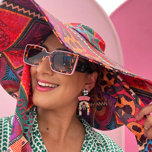 Close-up lifestyle view of a model wearing Tribe and Temple Confetti Multi Mixed Media Earrings. The geometric statement earring is styled with tortoise and pink square sunglasses, a vibrant patterned sun hat, and a green patterned shirt against a pink backdrop.