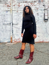 Medium front portrait of model Sarah wearing the black NOOWORKS Demi Dress outdoors against a white painted brick wall.