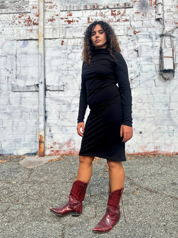 Medium front portrait of model Sarah wearing the black NOOWORKS Demi Dress outdoors against a white painted brick wall.
