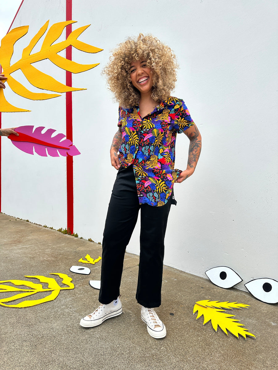 Model wearing Nooworks high-waisted Painter Pants in black twill paired with a colorful printed button-down shirt and white sneakers.