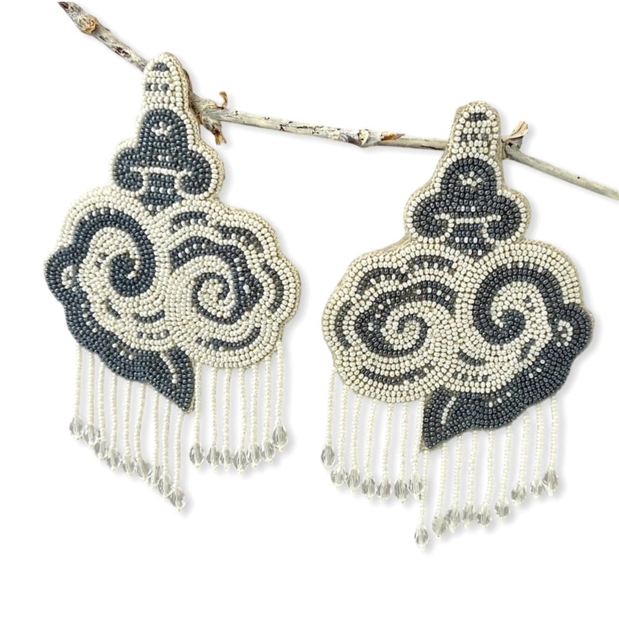 Pair of cloud-shaped hand-embroidered grey and white beaded statement earrings hanging from a tree branch.