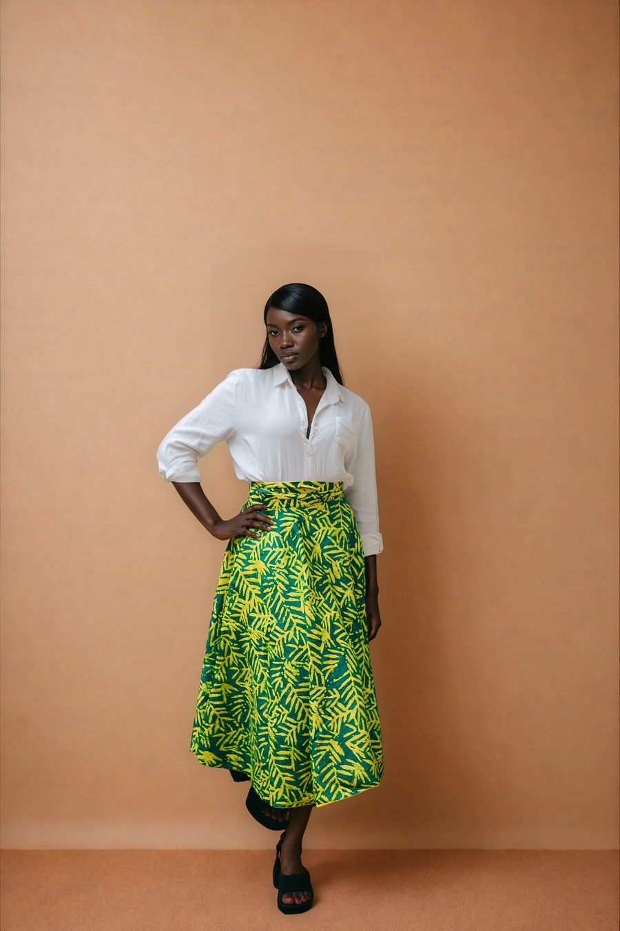 Full-length view of a model wearing the Besida Yellow Palm Reversible Wrap Skirt, showcasing the bold, hand-dyed green and yellow palm-leaf African print side. The high-waisted, mid-length wrap skirt is paired with a classic white long-sleeve button-down shirt and black open-toe platform sandals against a solid neutral beige background.