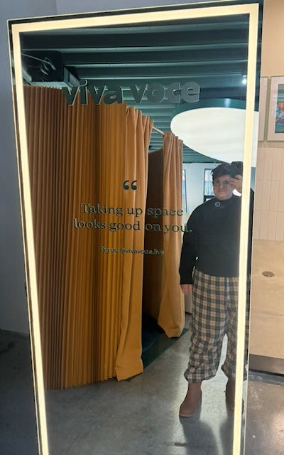 Kate Zigrang standing in front of a Viva Voce mirror with a reflection showing a room with a yellow partition.
