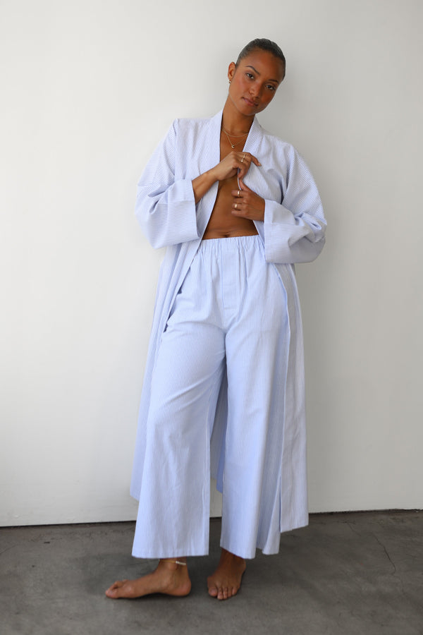 Full-body shot of a model standing in a size-inclusive light blue and white pinstripe lounge set by Be Idle, featuring the wide-leg Pinstripe Boxer Pant paired with a matching open robe.