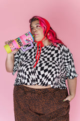 Medium studio portrait of model Angelina wearing the Nettle Studios Dizzy Favorite Tee in black and white wiggly checker plissé knit, holding a decorated tumbler against a pink backdrop.