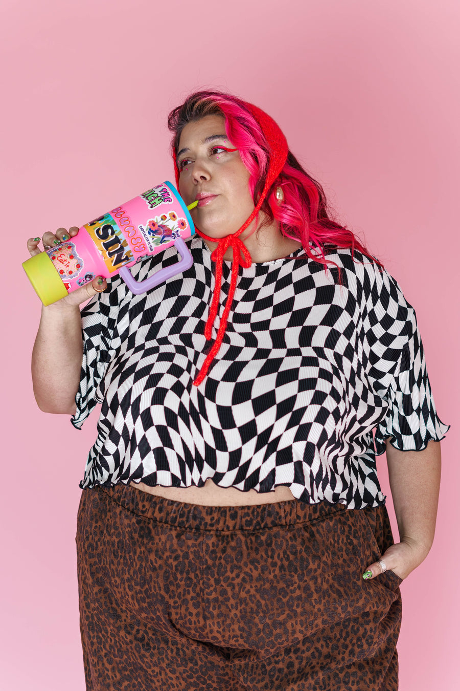 Medium studio portrait of model Angelina wearing the Nettle Studios Dizzy Favorite Tee in black and white wiggly checker plissé knit, holding a decorated tumbler against a pink backdrop.