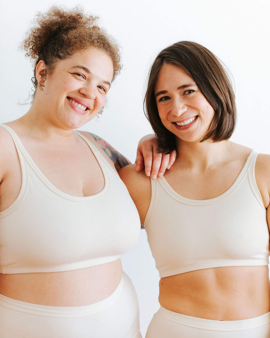 Close-up portrait of two smiling models standing shoulder to shoulder wearing matching cream organic cotton longline bra sets.
