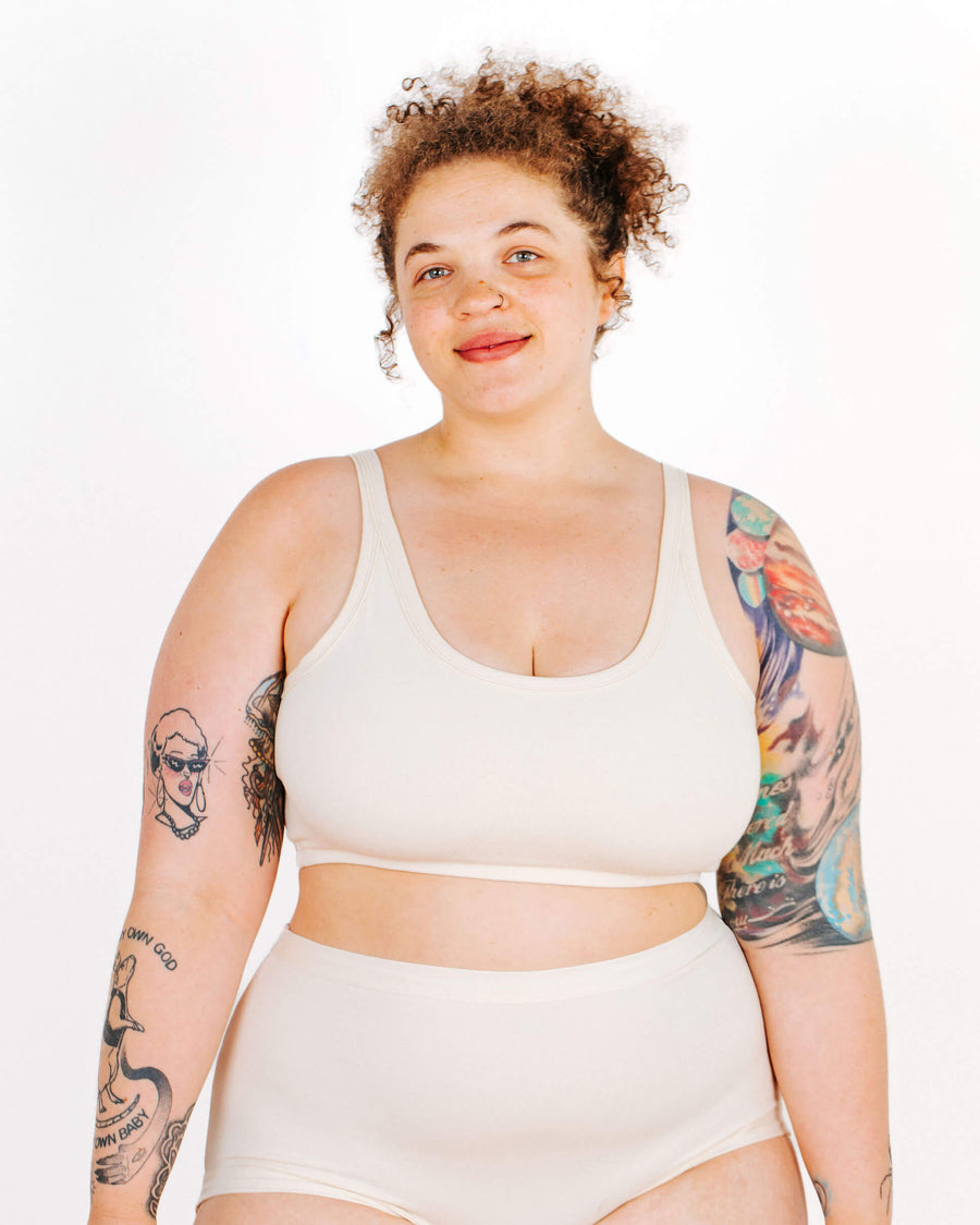 Front view portrait of a curly-haired model with arm tattoos wearing a cream organic cotton longline bra set.