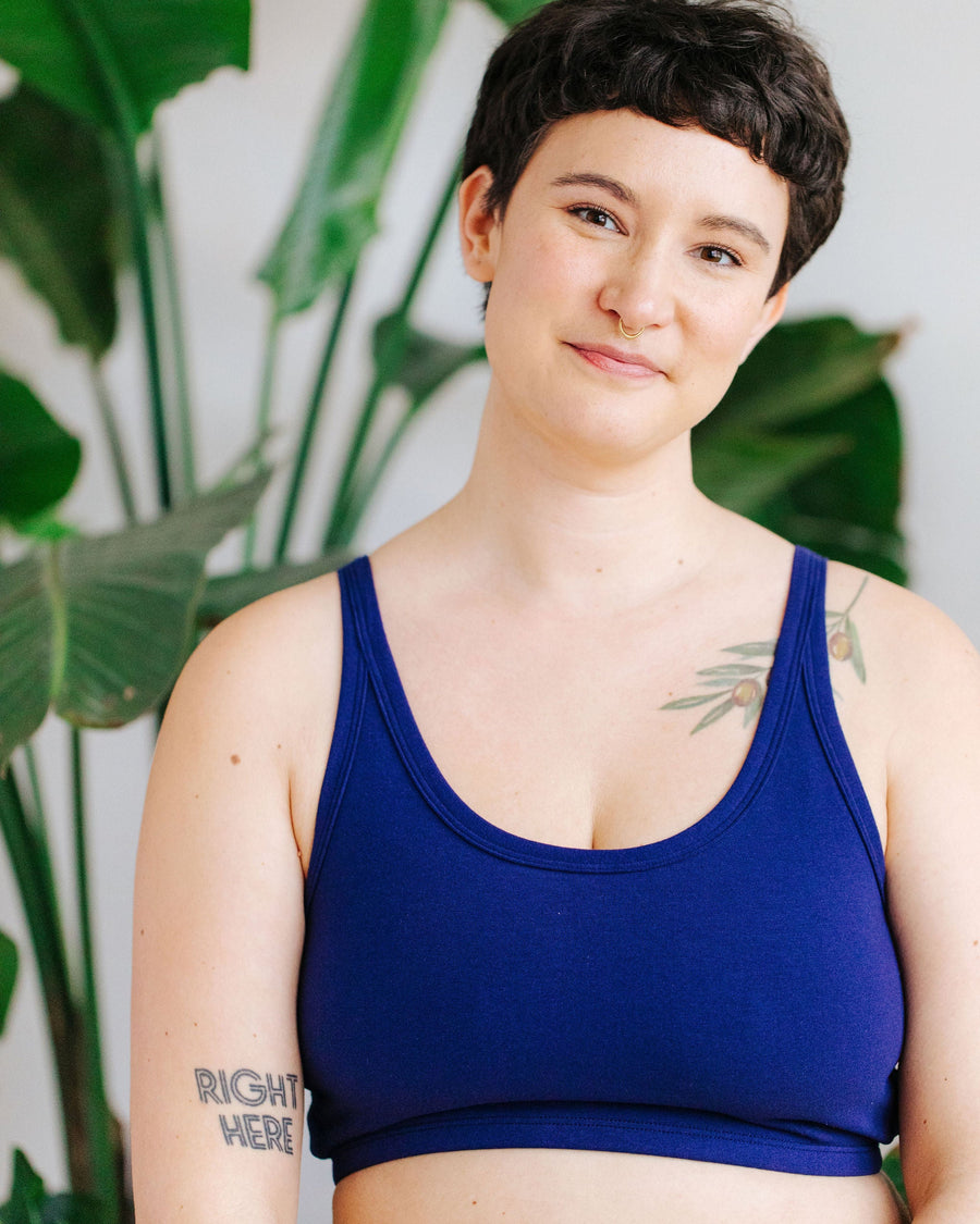 Close-up front view of a model wearing the organic cotton Indigo Montoya longline bra in deep blue against a backdrop of leafy green plants.