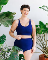 Three-quarter studio portrait of a model with short dark hair standing among houseplants wearing the Thunderpants US Indigo Montoya longline bra and matching deep blue high-waisted briefs.