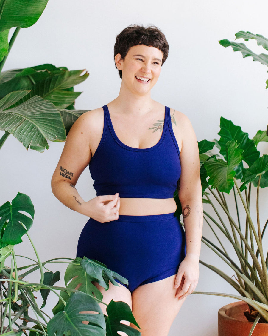 Three-quarter studio portrait of a model with short dark hair standing among houseplants wearing the Thunderpants US Indigo Montoya longline bra and matching deep blue high-waisted briefs.